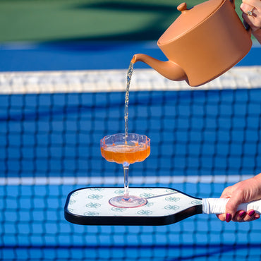 Tea being poured from a teapot into a glass on a paddle with a pickleball court in the background