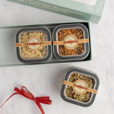 Three square metal tins of spices blends arranged on a light marble surface, with one tin open in a pale green gift box and another closed below it. A red ribbon is tied to the lower tin.
