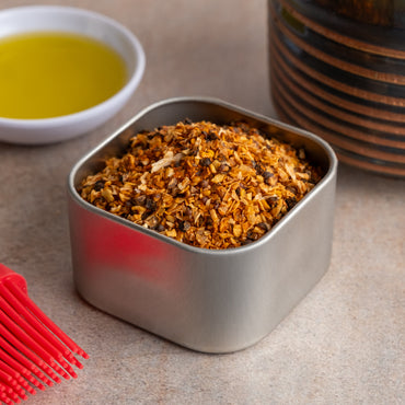 A metal container of Backwoods Hickory Rub Seasoning, filled with coarse orange-brown mix, sits on the countertop next to a bowl of olive oil, a red basting brush, and a striped ceramic jar.