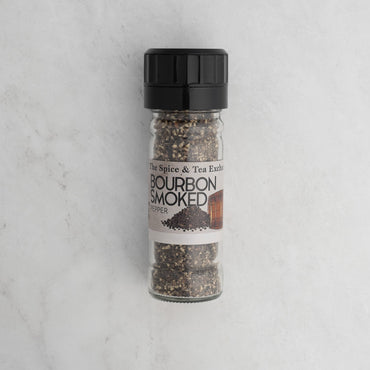 A clear grinder filled with Bourbon Smoked Black Pepper sits upright on a light marble surface.