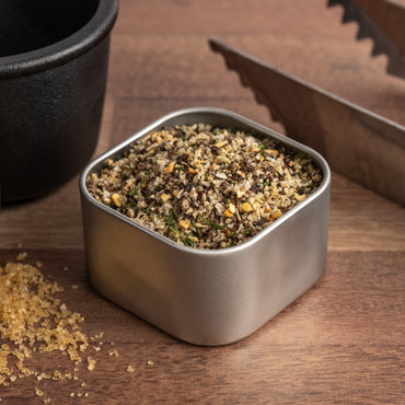 A square metal container of Butcher's Rub steak seasoning sits on a wooden surface beside tongs and a black bowl, with some coarse granules of the grilling spices scattered nearby.