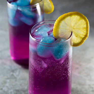 Two tall glasses of purple iced beverage with blue ice cubes, garnished with lemon slices, on a gray surface.