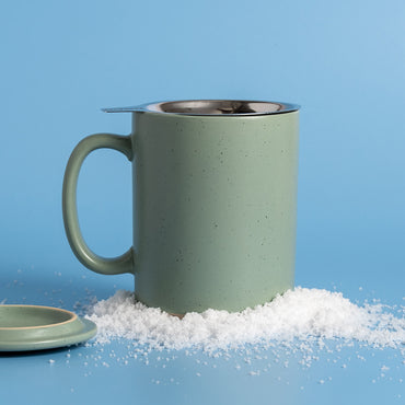 A speckled sage green ceramic mug with a stainless steel infuser lid, styled on a blue background with white snow crystals and a matching ceramic lid.