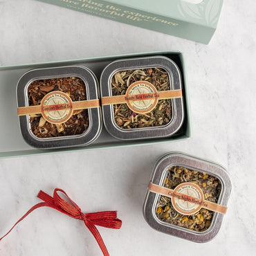 Three square metal tins of loose herbal tea displayed on a light marble surface, with one tin inside an open pale green gift box and the other placed outside the box. Each tin has a round label reading 'Herbal Tea' and is wrapped with a tan ribbon.