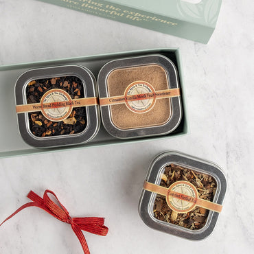 Three square metal tins of loose-leaf herbal tea displayed in a light green gift box, with a third tin tied with a red ribbon placed below on a light marble surface.