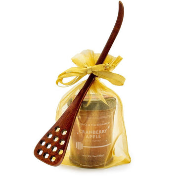 A Honey Jar with Dipper - Cranberry Apple Jam is placed in a sheer yellow gift bag, with a honeycomb wood dipper featuring hexagonal holes leaning against it.