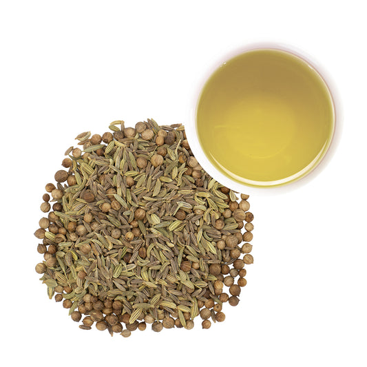 A pile of dried coriander cumin and fennel seeds next to a small white cup filled with pale yellow brewed tea, photographed on a clean white background.