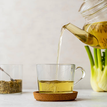 Clear glass teapot pouring pale yellow tea into a clear glass mug on a cork coaster, with a softly blurred kitchen-style background and green onions.
