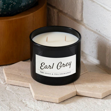 The Earl Grey Candle, a black natural soy jar from The Spice & Tea Exchange and inspired by Earl Grey Black Tea, sits on beige hex tiles by a white brick wall and wood surface. The jar features two unlit wicks.