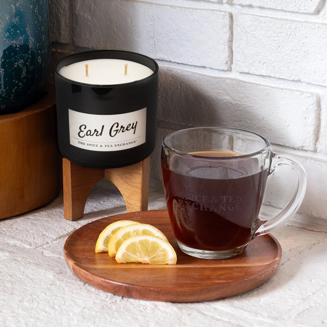 A glass mug of Earl Grey Black Tea sits on a wooden plate with three lemon slices. Nearby, a bergamot-scented candle in a black holder is displayed against a white brick wall.