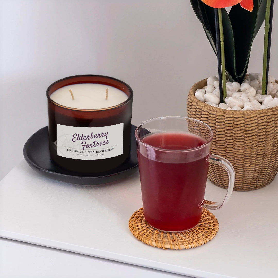Candle labeled 'Elderberry Fortress' and a mug of red tea on a white surface with a plant in the background.