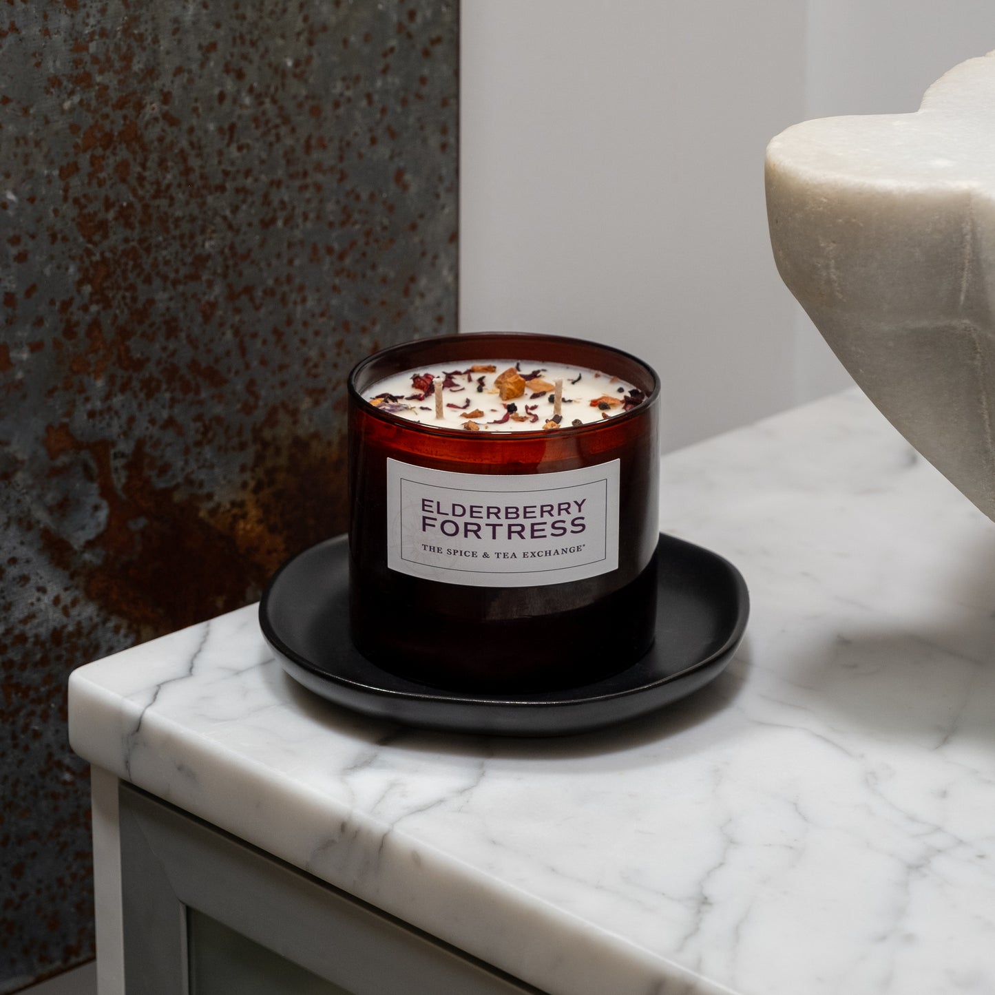 The Elderberry Fortress Candle in brown glass, crafted from natural soy and essential oils, sits on a black saucer atop a white marble countertop beside a large stone decor piece and a rusty metal background.