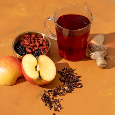 Elderberry Fortress Herbal Tea brewed in glass mug next to elderberries, hibiscus, apple slices, ginger root, and goji berries