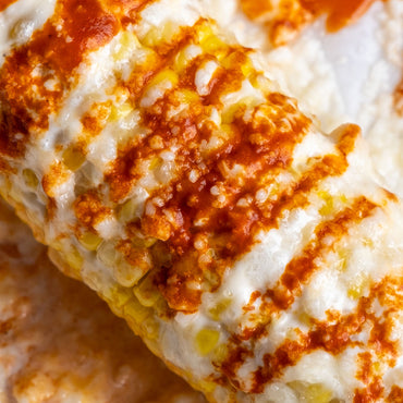 Close-up of corn topped with melted white cheese and topped with orange-red sauce, showing corn kernels and a crispy textured surface.