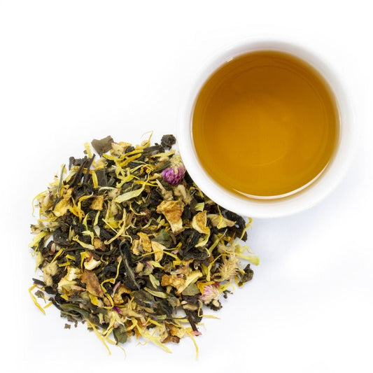 A white cup of Florida Sunshine White Tea sits beside a pile of loose leaf tea with green leaves, yellow petals, and dried flower buds on a white background.