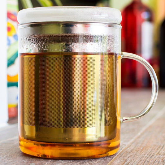 The Glass Tea Mug Infuser, made of clear heat-resistant glass with a stainless steel infuser, sits on a wooden surface filled with steaming amber tea. A softly blurred background adds a cozy touch.