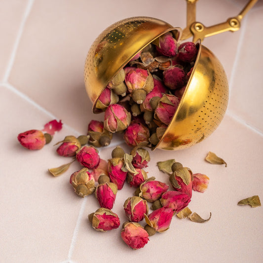 The Gold Scissor Infuser, filled with dried pink rosebuds, rests on a pale tiled surface, with some rosebuds nestled inside and others scattered around it.