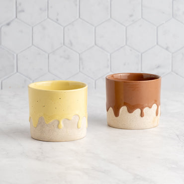 Two ceramic cups, one yellow and one brown, on a light gray surface with a hexagonal tile pattern.