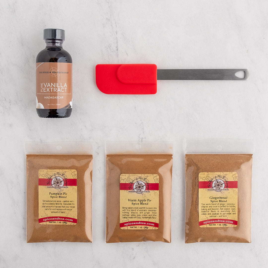 A flat-lay featuring a bottle of pure vanilla extract, a red silicone spatula, and three bags of spice blends.