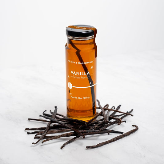 A 12 oz Vanilla Honey Jar from The Spice & Tea Exchange sits on a white surface, surrounded by dried vanilla beans, highlighting the rich blend of raw honey and aromatic vanilla.