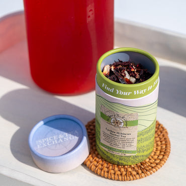 Brewed red tea in pitcher next to white decorative tube of Lychee Hibiscus Herbal Tea