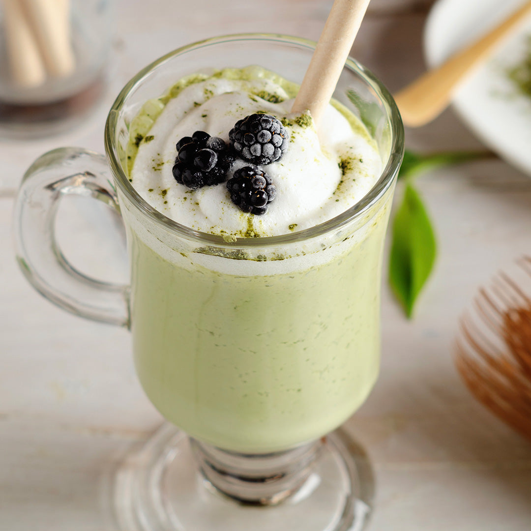 A clear glass mug filled with a light green beverage, topped with a thick white whipped cream or foam layer, fresh blackberries, and a wooden straw, photographed on a light tabletop with softly blurred background props.