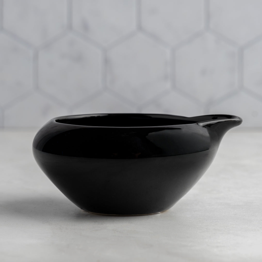 Black ceramic bowl with a handle on a light gray surface with a geometric patterned wall in the background.