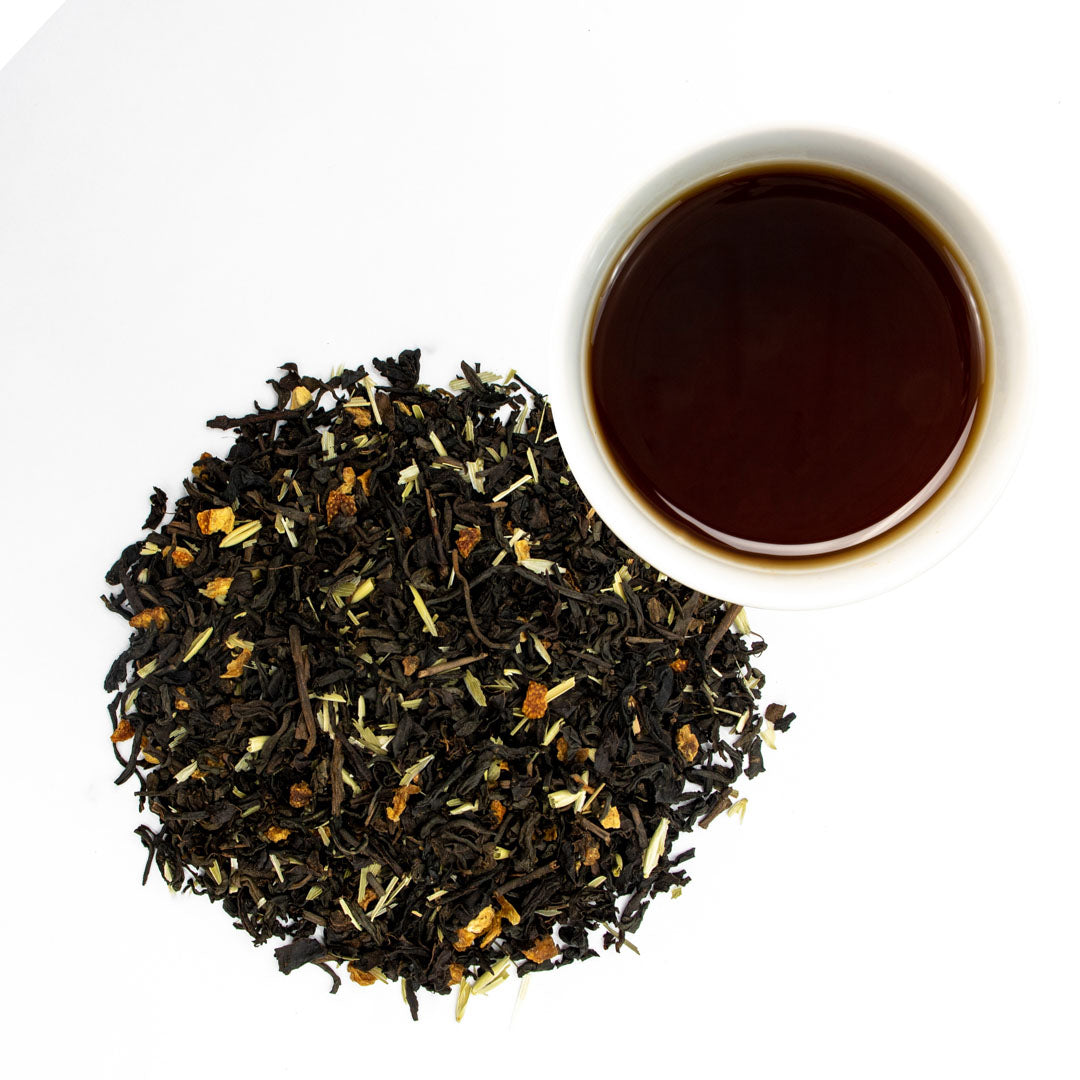 A pile of Morning Mindset Pu-erh Black Tea with orange and yellow pieces sits next to a white cup of dark brewed tea on a white background.