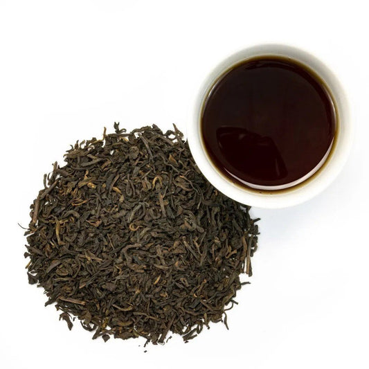 A pile of Organic Black Pu-erh Tea with an earthy aroma sits beside a white cup of brewed dark tea, all displayed on a white background.