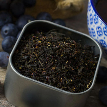 A metal tin of Blueberry Black Tea sits on a wooden surface, next to a blue-patterned teacup and fresh blueberries.