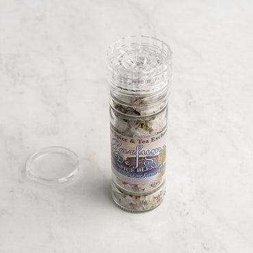 A clear spice grinder labeled Parfume de Sel Spice Blend from The Spice & Tea Exchange sits on a light marble surface, with its detached cap resting beside the bottle.