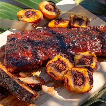 Grilled barbecue ribs on a wooden board, surrounded by halved, grilled peaches with char marks. The scene suggests a summer barbecue setting.