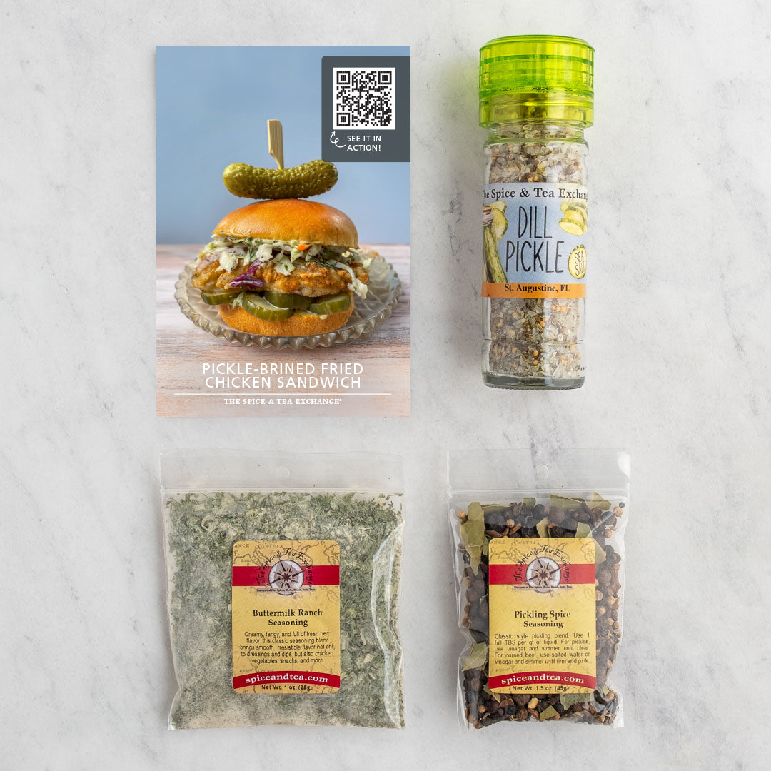 A flat-lay of a spice and tea seasoning gift set on a white marble background, featuring a pickle-brined fried chicken sandwich recipe card, a glass bottle of dill pickle with a green cap, and two clear bags of seasoning blends.