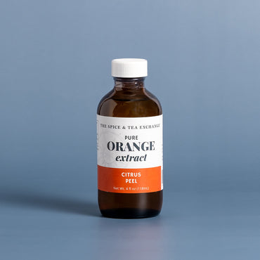 A bottle of Tree Spice & Tea Exchange Pure Orange Extract (Citrus Peel) in an amber glass bottle with a white cap and white label featuring an orange accent band, set against a solid blue-gray background.