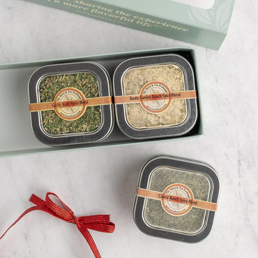 A gift set of three square metal tins of spice blends arranged in an open pale green box on a light marble surface, with one tin placed below and a red ribbon setting next to it. The tins have round labels.