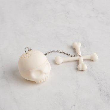 An off-white skull-shaped container with a small silver hanging loop, connected by a short silver chain to an off-white cross-bone charm, photographed on a light marble surface.