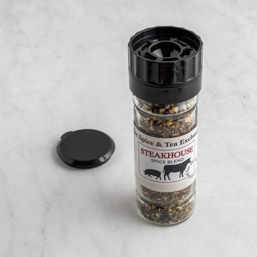 A clear spice blend shaker bottle labeled 'Steakhouse Spice Blend' by The Spice & Tea Exchange, with a black cap and lid placed beside it on a light marble surface.