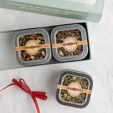 A gift set of herbal tea tins displayed on a light marble surface, with the open box lid at the top and a red ribbon sitting next to it. The set includes two square silver tins in the box labeled Herbal Tea and one single tin below labeled Herbal Tea.