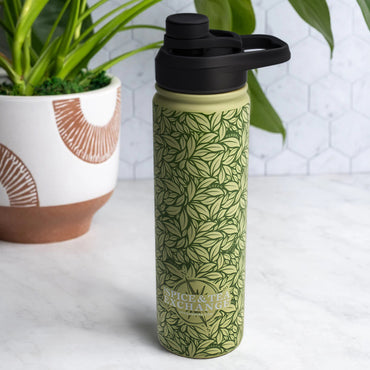 The Tea Leaf Travel Infuser Tumbler, a green stainless steel mug with a leafy pattern and Spice & Tea Exchange logo, sits on white marble beside a potted plant and against a white hexagonal tile background.