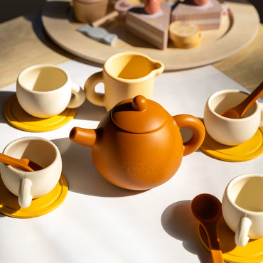 The Tiny Tea Party Toy Set features a brown children’s tea set with four cream and yellow teacups, matching saucers, and spoons. Toy desserts on a wooden platter complete this perfect pretend play setup under soft sunlight.