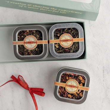 Two square metal tins of loose-leaf herbal tea displayed on a light marble surface, with one tin inside an open pale green gift box and the other placed below. Each tin has a tan paper band and a round label.