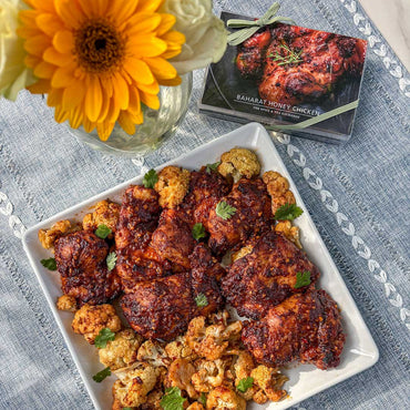 A square white plate holds Baharat Honey Chicken (Kit) with roasted chicken and cauliflower, seasoned with Baharat Spice Blend and garnished with herbs. It sits on a blue placemat beside a yellow flower and the Baharat Honey Chicken meal box.