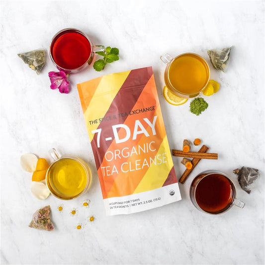 A 7 Day Organic Tea Cleanse package is displayed with four cups of colorful organic tea, tea bags, flower petals, lemon slices, turmeric, cinnamon sticks, and loose tea leaves on a white background.