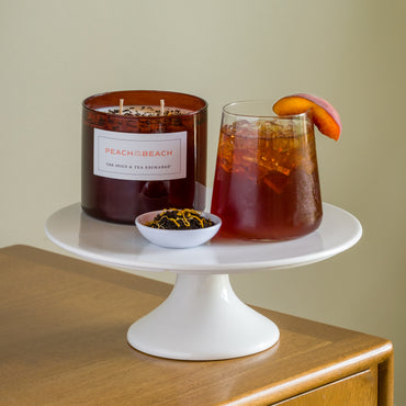 A glass of iced tea with a peach slice, the Peach on the Beach Candle, and a bowl of loose tea leaves are arranged on a white cake stand on a wooden surface.