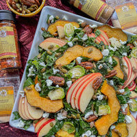 A square white plate holds a vibrant salad with kale, apple slices, roasted squash, pecans, pumpkin seeds, goat cheese, and Brussels sprouts, surrounded by spice jars and a small bowl of seeds on a red tablecloth.
