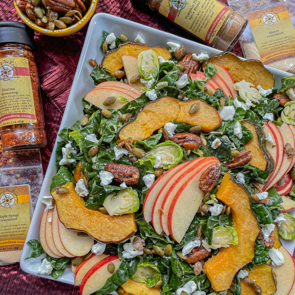 A square white plate holds a vibrant salad with kale, apple slices, roasted squash, pecans, pumpkin seeds, goat cheese, and Brussels sprouts, surrounded by spice jars and a small bowl of seeds on a red tablecloth.