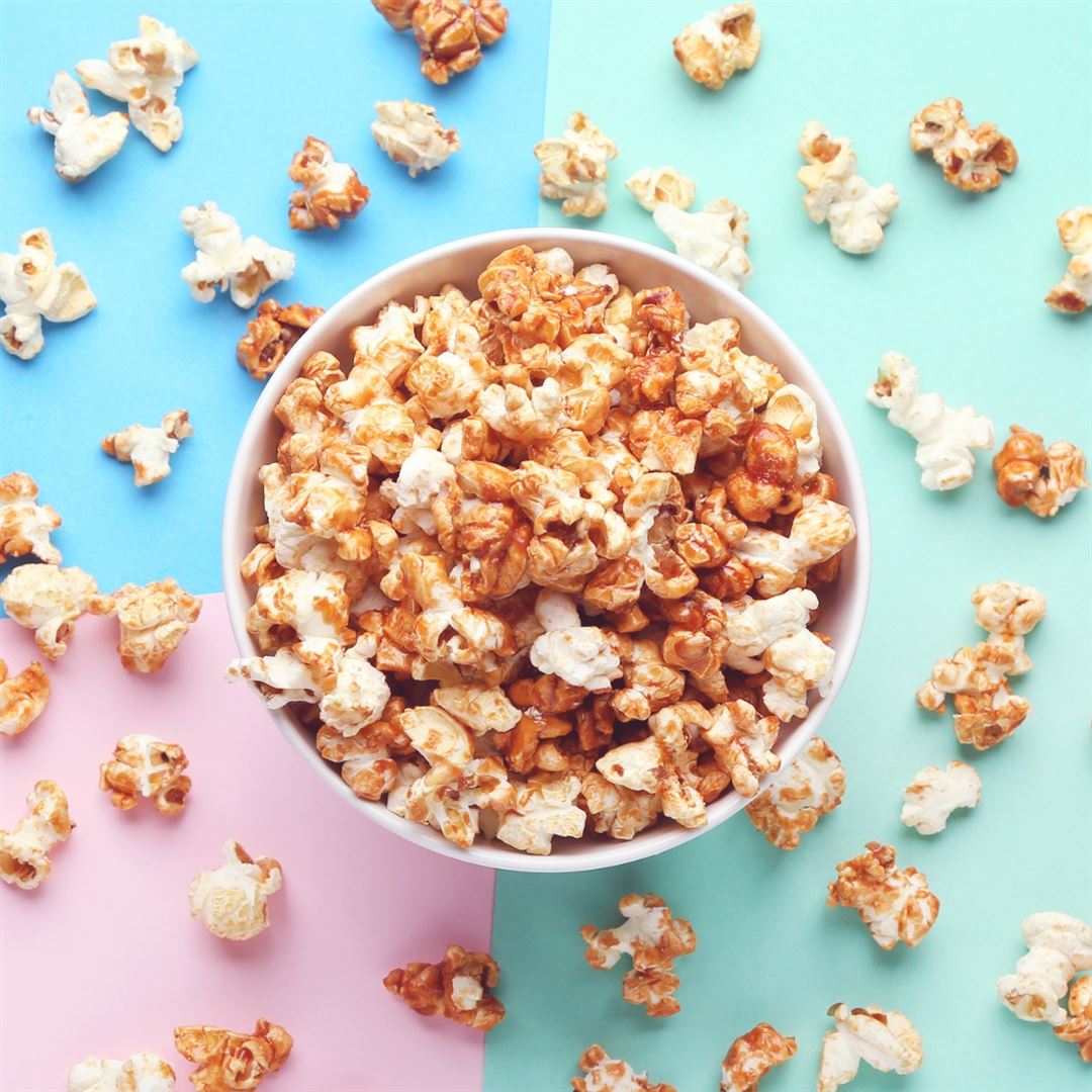 A bowl of caramel and plain popcorn sits on a pastel tri-colored background with blue, green, and pink sections. Several popcorn pieces are scattered around the bowl.