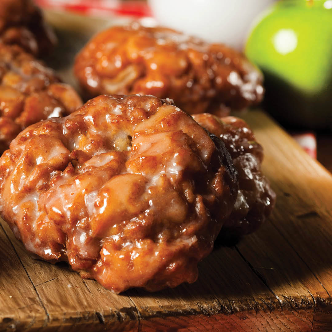 Close-up of glazed apple fritters with a shiny, caramelized exterior, resting on a rustic wooden board. A green apple is slightly out of focus in the background.