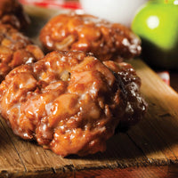 Close-up of glazed apple fritters with a shiny, caramelized exterior, resting on a rustic wooden board. A green apple is slightly out of focus in the background.