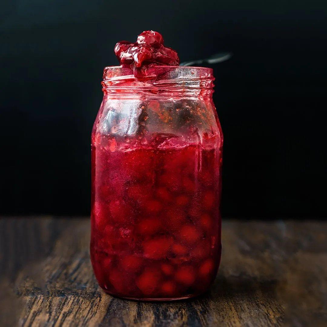A glass jar filled with bright red cherry preserves sits on a wooden surface, with a spoon resting on top and some preserves dripping over the jar’s rim against a dark background.